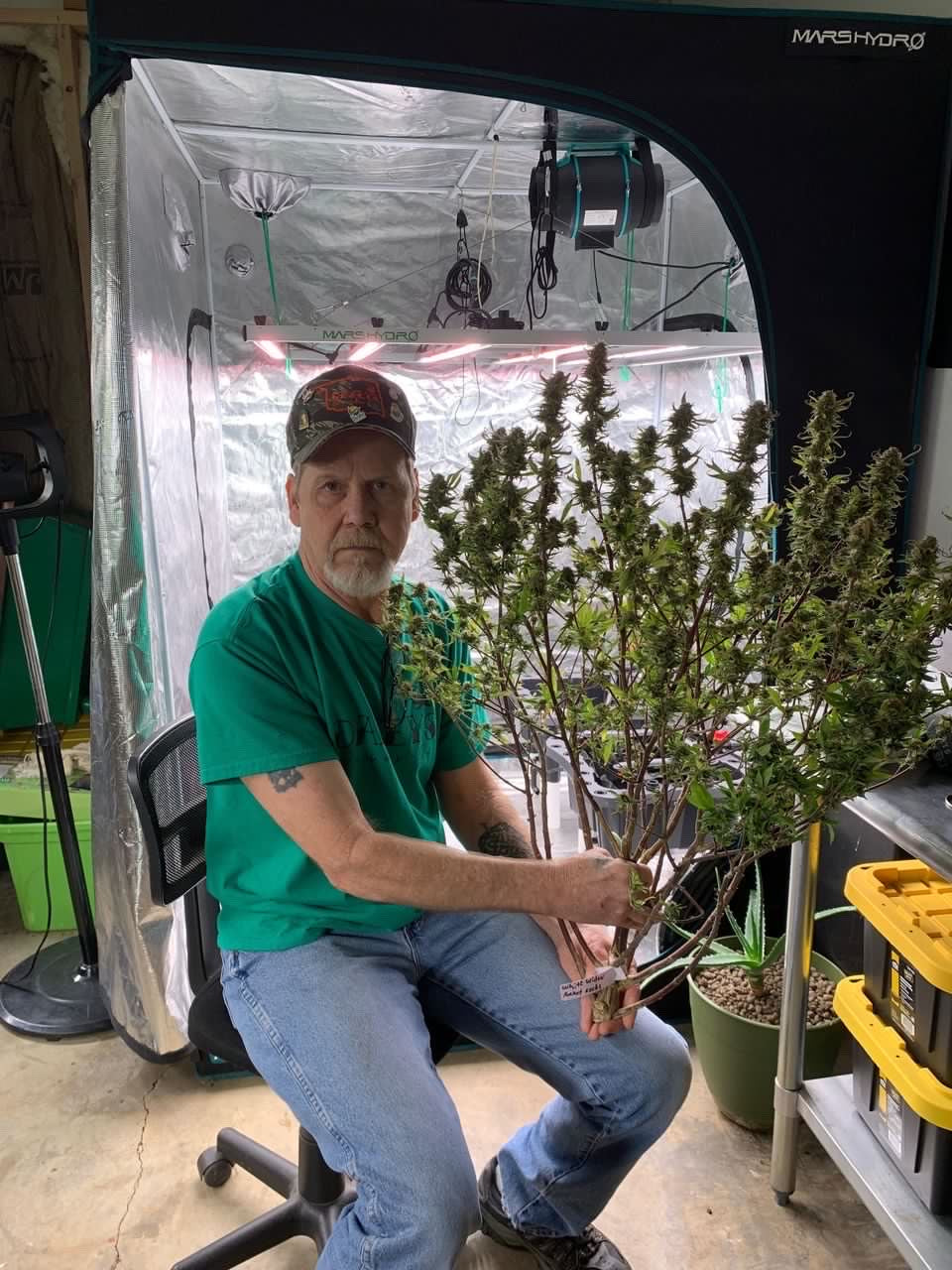 Man sitting in a grow room holding a large cannabis plant, shared by @heavlymedicated. The testimonial describes a positive experience with Rhize Nutrients, praising the ease of use, support through charts and videos, and achieving strong plant health without extra additives.